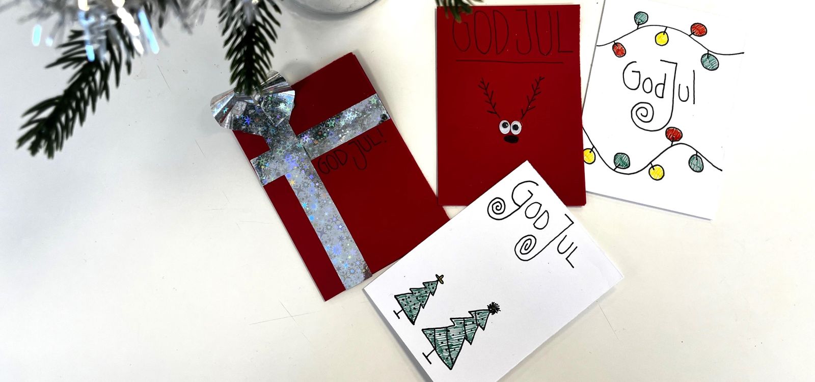 Handgjorda julkort med texten God Jul dekorerade med glitterband, renmotiv, julgranar och ljusslingor, placerade på vitt underlag med en grankvist i hörnet.